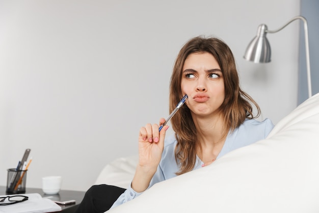 Imagem de uma mulher bonita jovem bonita pensativa em casa dentro de casa escrevendo notas no caderno.