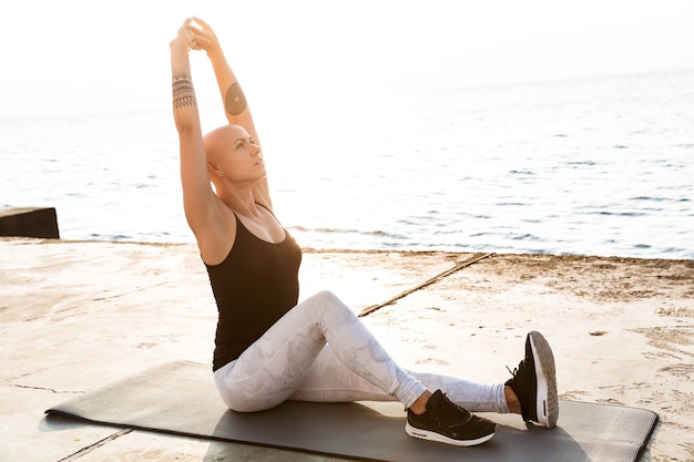 Imagem de uma mulher calva e calva em roupas esportivas fazendo exercícios no colchonete enquanto se exercita perto da beira-mar em uma manhã ensolarada