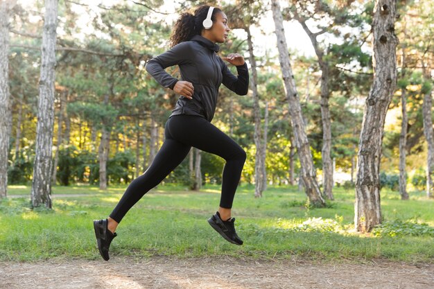 Imagem de uma mulher feminina com 20 anos, vestindo um agasalho esportivo preto e fones de ouvido, malhando, enquanto corre pelo parque