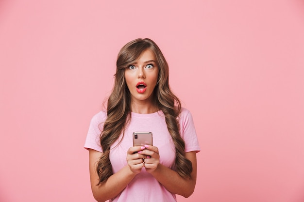 Imagem de uma mulher morena surpresa com um lindo cabelo comprido, olhando para você com olhos esbugalhados, segurando o celular nas mãos, isolado sobre um fundo rosa