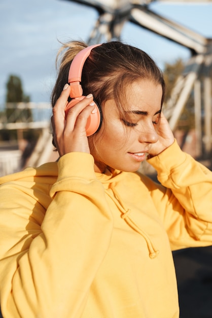 Imagem de uma mulher sorridente de fitness muito jovem posando ao ar livre, ouvindo música com fones de ouvido.