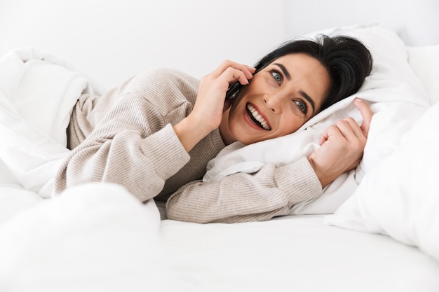 Imagem de uma mulher sorridente na casa dos 30 anos usando smartphone, deitada na cama com lençóis brancos em casa