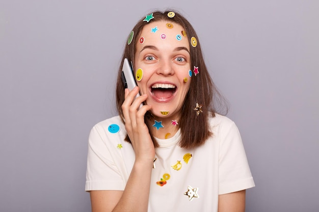 Imagem de uma mulher surpresa espantada coberta com adesivos engraçados posando isolada sobre fundo cinza falando ao telefone ouvindo notícias agradáveis regozijando-se olhando para a câmera