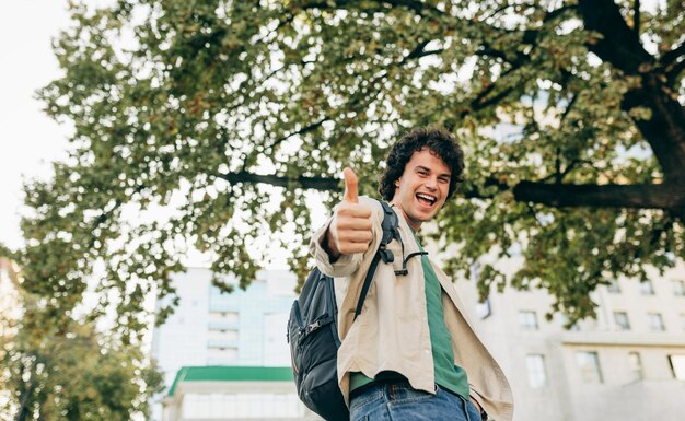 Imagem de vista inferior de homem sorridente bem sucedido com cabelo encaracolado posando para propaganda social com o polegar para cima posando na rua da cidade Macho de estudante alegre animado fazendo um bom gesto