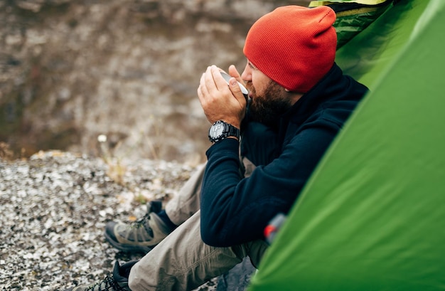 Imagem de vista lateral do jovem explorador bebendo bebida quente nas montanhas sentado perto da fogueira relaxando depois de trekking Homem viajante de chapéu vermelho segurando uma caneca de chá depois de caminhar Pessoas de viagem