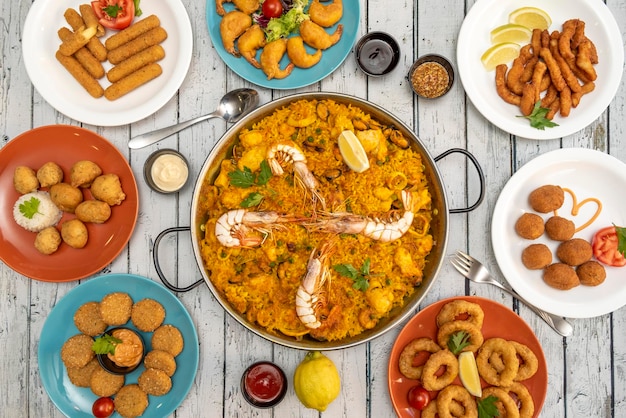 Imagem de vista superior de pratos de aperitivo e uma paella de frutos do mar no centro sobre uma mesa de madeira com talheres e molhos