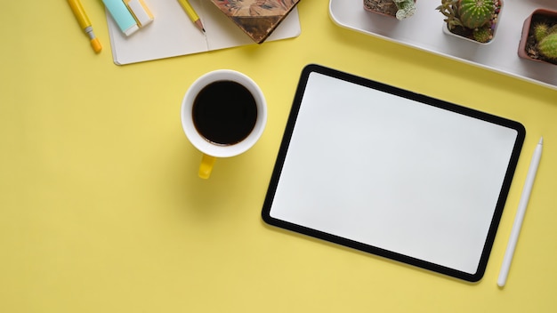 Imagem de vista superior do local de trabalho colorido. Lápis bússola, xícara de café, caderno, canetas, cacto, caneta stylus e tablet computador com tela vazia branca plana leigos na mesa amarela.