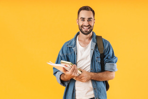 Foto imagem do aluno de conteúdo em roupas jeans sorrindo enquanto segura cadernos isolados sobre a parede amarela