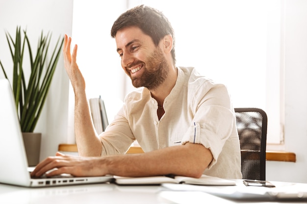 Imagem do empresário atraente dos anos 30, vestindo uma camisa branca, trabalhando com laptop e documentos em papel, enquanto está sentado em um escritório bem iluminado