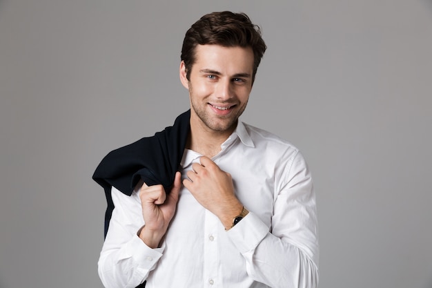 Imagem do empresário positivo dos anos 30 sorrindo e segurando uma jaqueta por cima do ombro, isolada sobre uma parede cinza