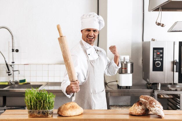 Imagem do homem alegre padeiro em uniforme branco sorrindo, enquanto estava na padaria com pão na mesa