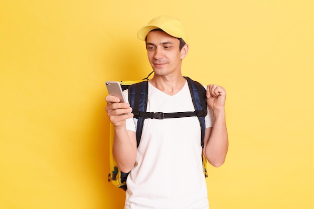 Imagem do homem de correio concentrado calmo de boné amarelo e camiseta segurando o telefone inteligente e olhando para a tela encontrando o endereço do cliente navegando na internet durante o intervalo isolado no fundo amarelo