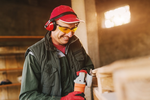 Imagem do homem maduro de sorriso do carpinteiro no uniforme protetor que dá forma à madeira com moedor elétrico. Apreciando seu trabalho em sua garagem em dia ensolarado.
