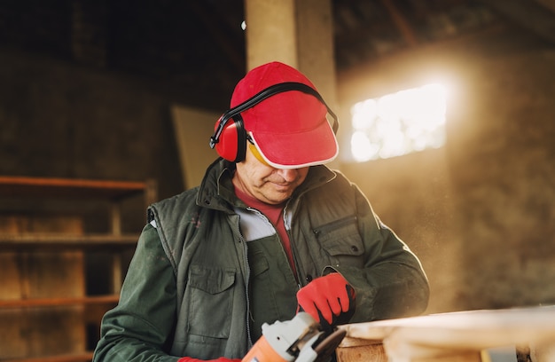 Imagem do homem maduro do carpinteiro no uniforme protetor que dá forma à madeira com moedor elétrico. Apreciando seu trabalho em sua garagem em dia ensolarado.