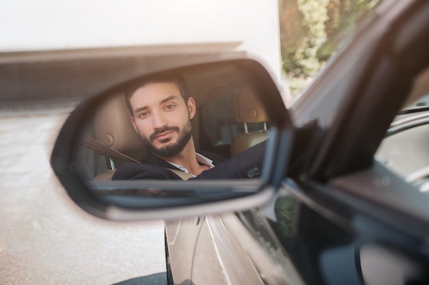 Imagem do homem sentado no carro e olhando para o espelho de asa. Ele sils um pouco. Jovem satisfeito com seu carro. Está ensolarado lá fora.