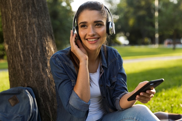 Imagem do incrível estudante de mulher jovem e bonita no parque ouvindo música com fones de ouvido usando telefone celular.