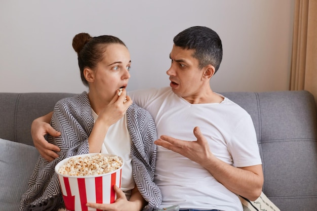 Imagem do jovem casal sentado no sofá e assistindo filme mulher envolta em cobertor comendo pipoca marido olhando para ela com expressão chocada e confusa