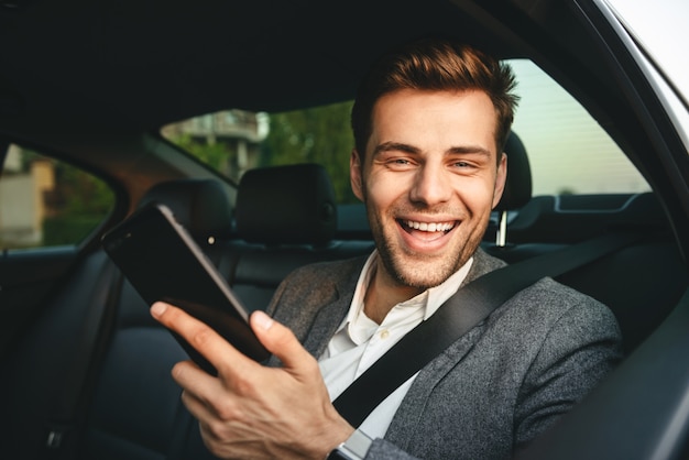 Imagem do jovem diretor homem de terno segurando o smartphone e sorrindo, enquanto volta sentado no carro de classe executiva com cinto de segurança