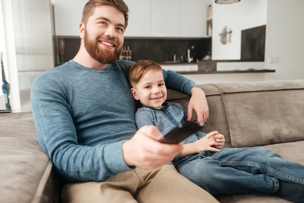 Imagem do jovem pai barbudo segurando o controle remoto enquanto assiste tv com seu filho pequeno e bonito.