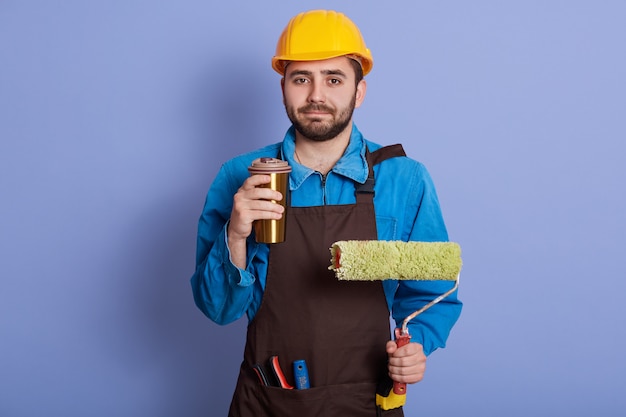 Imagem do reparador agradável de trabalho duro que guarda a caneca thermo com bebida quente e rolo para a pintura de parede, tendo a pausa para o café, vestindo uniforme com equipamento, tendo o descanso, apreciando a hora do almoço.