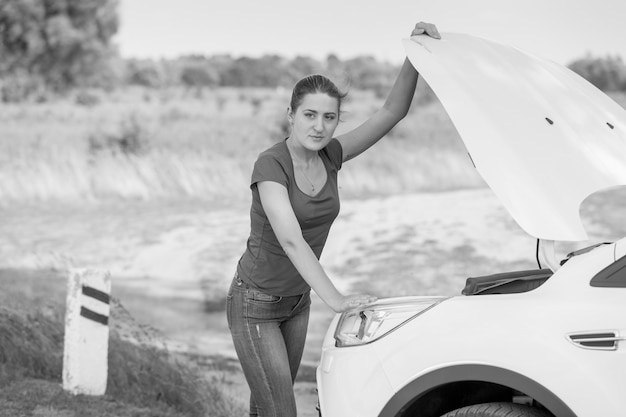 Imagem em preto e branco de uma jovem abrindo o capô de um carro quebrado na beira da estrada