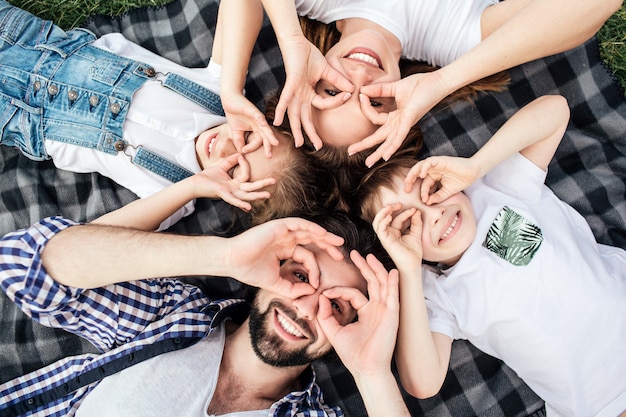 Imagem engraçada da família fazendo rodadas engraçadas com os dedos nos olhos. eles estão jogando. todos eles estão deitados no cobertor e sorrindo. eles parecem felizes.