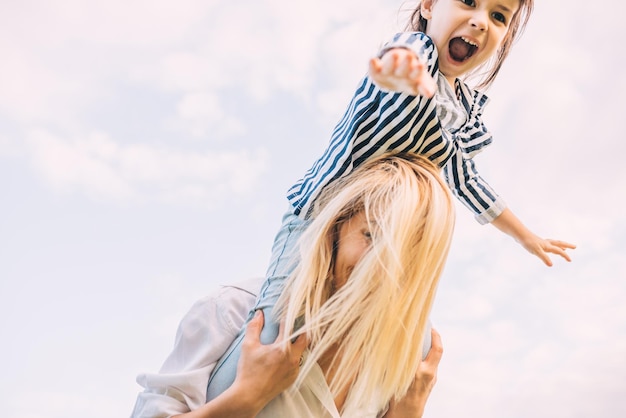 Imagem horizontal de filha feliz em um passeio nas costas com sua mãe sorridente no fundo do céu Mulher amorosa e sua filha brincando no parque Mãe e filho se divertem fora da maternidade