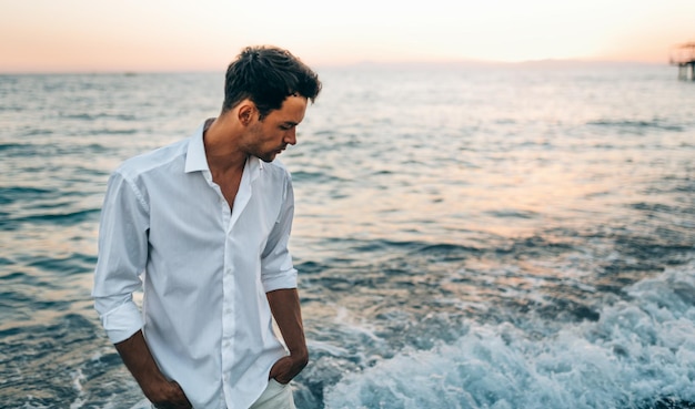 Imagem horizontal de homem bonito, vestindo camisa branca no fundo do mar no susnet Viagem férias férias Macho atraente andando na praia do oceano aproveite a temporada tropical