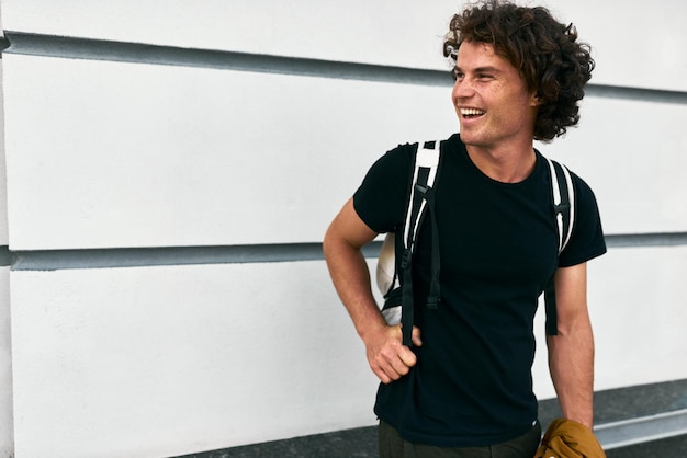 Imagem horizontal de homem sorridente bonito com cabelo encaracolado com mochila em pé na parede de construção na rua enquanto espera seus colegas Empresário usa camiseta preta casual Copie o espaço para texto