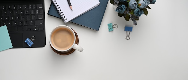 Imagem horizontal de mesa de escritório branca com xícara de café, planta, caderno e teclado.