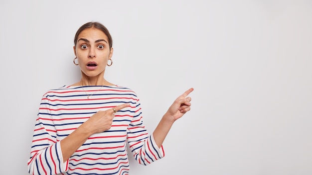 Imagem horizontal de mulher impressionada fala sobre a descoberta inacreditável indica no canto superior direito suspiros espantados usando macacão listrado casual olhando em estupor isolado sobre fundo branco