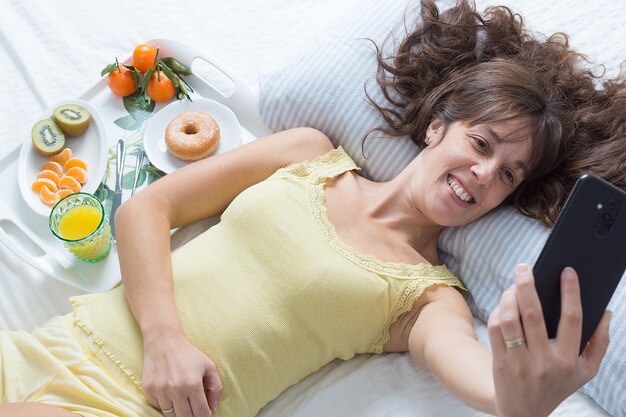 Imagem horizontal de uma jovem sorridente de cabelos castanhos deitada na cama com uma camiseta amarela tirando uma selfie ao lado de uma bandeja de café da manhã na luz suave da manhã