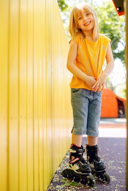 Foto imagem interior de uma menina brincalhona, enérgica e ativa, levantando as mãos, sentada no chão, isolada sobre um fundo amarelo, vestindo roupas casuais cor-de-rosa e patins, sorrindo sinceramente.