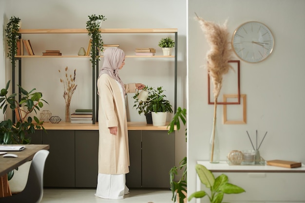 Imagem longa de mulher muçulmana irreconhecível vestindo roupas confortáveis e hijab regando plantas de casa no espaço de cópia em casa