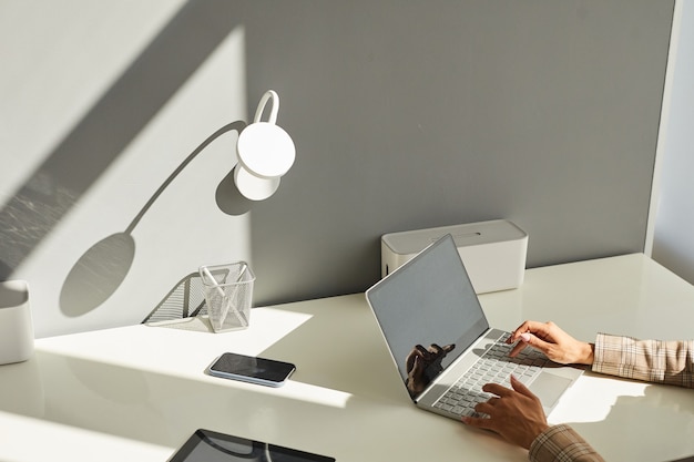 Imagem mínima da superfície de uma mulher irreconhecível usando o laptop na mesa branca do local de trabalho com foco nas elegantes mãos femininas digitando no teclado à luz do sol, copie o espaço