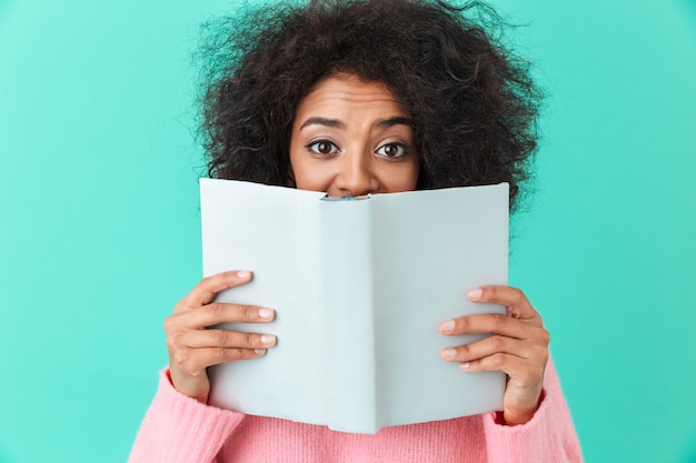 Imagem positiva da mulher americana 20 anos com penteado afro e rosto coberto com livro interessante, isolado sobre parede azul