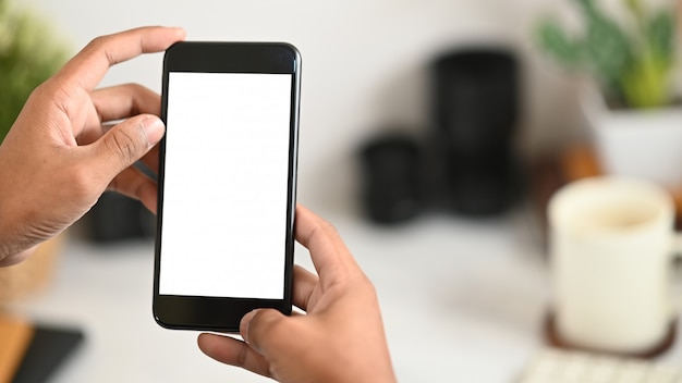 Foto imagem recortada das mãos do homem inteligente, segurando um smartphone preto recortado com tela em branco branca sobre sua mesa de trabalho branca.