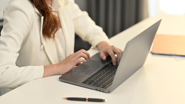 Imagem recortada de empresária em terno elegante branco sentado em sua mesa de trabalho e usando computador portátil