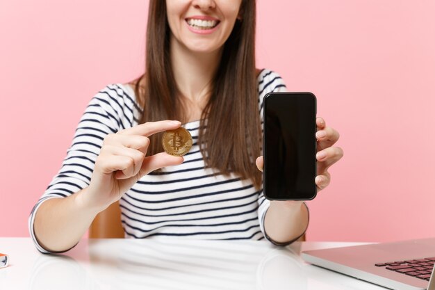 Imagem recortada de mulher segurando uma moeda de metal bitcoin de cor dourada, telefone celular de moeda futura com tela em branco vazia, sente-se na mesa