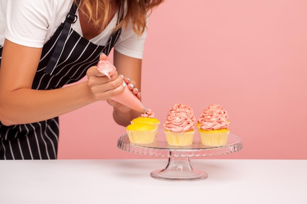 Foto imagem recortada de padaria mulher decorando muffins com creme rosado usando deliciosa sobremesa de cone de pastelaria na placa de vidro confeiteiro sem rosto trabalhando estúdio interno tiro isolado no fundo rosa