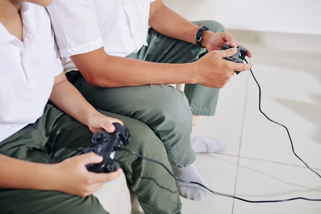 Imagem recortada de pai e filho sentados no sofá em casa, jogando videogame juntos