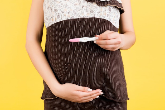 Imagem recortada de teste de gravidez positivo com duas listras contra o abdômen da mulher grávida em fundo amarelo futura mãe de pijama marrom surpresa de gravidez espaço de cópia