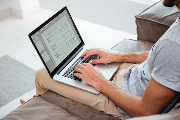 Imagem recortada de um jovem que trabalha em seu laptop enquanto está sentado no sofá.