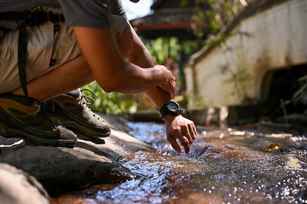 Imagem recortada de um trekker masculino asiático sentado perto do pequeno córrego da montanha tocando a água