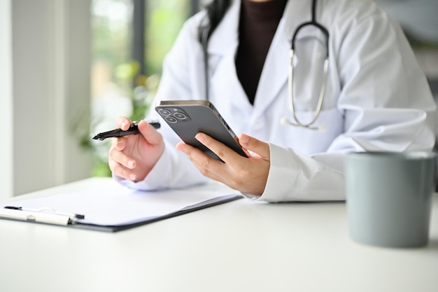 Imagem recortada de uma médica asiática inteligente usando seu smartphone em sua mesa de escritório