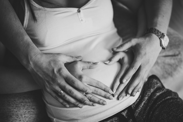 Imagem recortada do marido segurando a barriga da esposa grávida, fazendo as mãos do coração do símbolo. Mulher grávida e homem abraçando a barriga em casa. Conceito de paternidade. Chá de bebê. Foto em preto e branco.