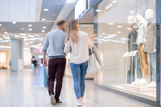 Imagem traseira de jovens compradores prestando atenção na nova coleção de roupas casuais masculinas na vitrine enquanto passam algum tempo no shopping