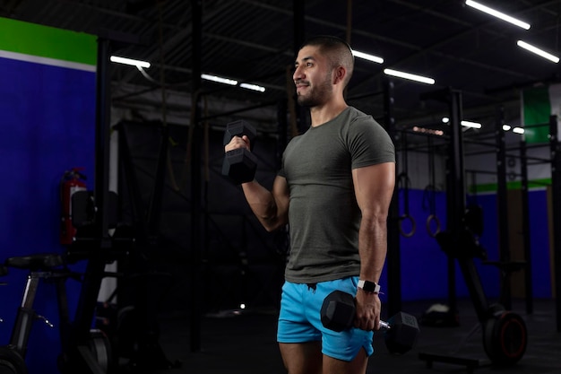 Imagem vertical de um atleta latino fazendo exercícios de bíceps com dois halteres em uma academia