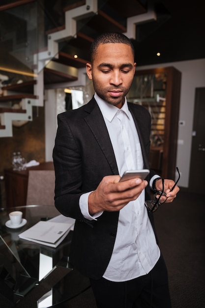 Imagem vertical de um homem africano sério de terno, usando o telefone no hotel