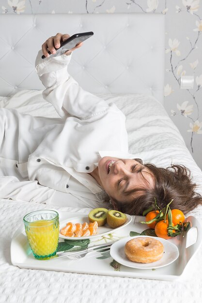 Imagem vertical de uma mulher deitada na cama com uma camisa branca tirando uma selfie ao lado de uma bandeja de café da manhã na cama na luz suave da manhã